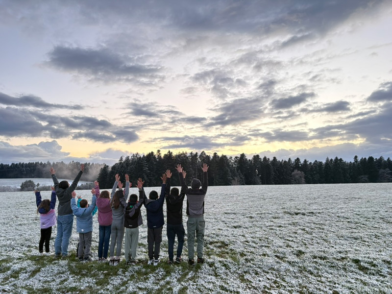 Gruppe von Kindern und Erzieher:innen in einer Winterlandschaft, alle strecken die Arme nach oben